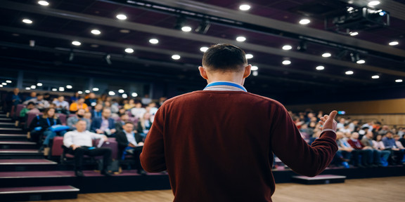 Foto: Ein Mann in braunem Pullover steht in einem vollen Konferenzsaal vor Publikum.