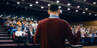 Foto: Ein Mann in braunem Pullover steht in einem vollen Konferenzsaal vor Publikum.