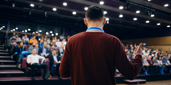 Photo: A man in a brown sweater stands in front of an audience in a crowded conference room.