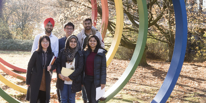 Group photo of six international students standing next to a sculpture of five colourful rings on Campus North