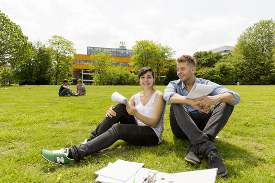 Zwei Studierende der TU Dortmund sitzen auf einer grünen Wiese. Im Hintergrund sieht man das gelbe Mensagebäude der TU Dortmund.