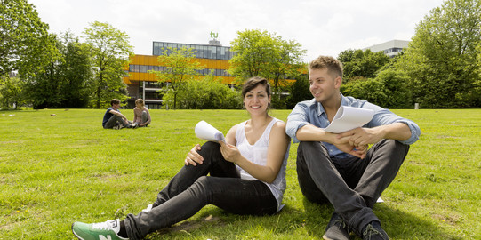 Zwei Studierende der TU Dortmund sitzen auf einer grünen Wiese. Im Hintergrund sieht man das gelbe Mensagebäude der TU Dortmund.