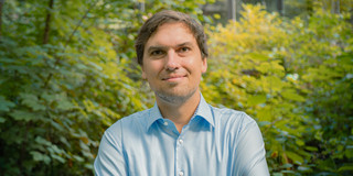 Portrait photo of Christian Glaser: Prof. Glaser is wearing a light blue shirt and has his arms crossed. There are bushes in the background.