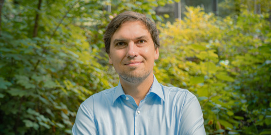 Portrait photo of Christian Glaser: Prof. Glaser is wearing a light blue shirt and has his arms crossed. There are bushes in the background.