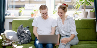 Im Zentrum des Bildes sind zwei junge Erwachsene zu sehen, die auf einem grünen Sofa sitzen. Beide tragen Jeans und ein helles Oberteil. Die männlich gelesene Person hält einen aufgeklappten, silbernen Laptop auf seinen Oberschenkeln. Beide Personen richten den Blick auf den Bildschirm. Im Hintergrund ist ein großes Fenster sowie eine Fensterbank mit einigen Topfpflanzen zu sehen.