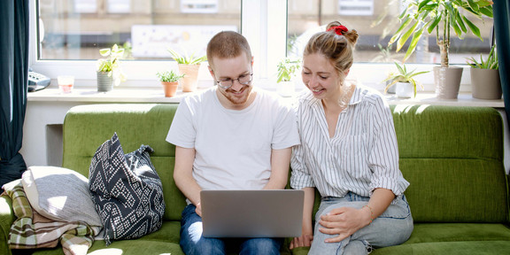 Im Zentrum des Bildes sind zwei junge Erwachsene zu sehen, die auf einem grünen Sofa sitzen. Beide tragen Jeans und ein helles Oberteil. Die männlich gelesene Person hält einen aufgeklappten, silbernen Laptop auf seinen Oberschenkeln. Beide Personen richten den Blick auf den Bildschirm. Im Hintergrund ist ein großes Fenster sowie eine Fensterbank mit einigen Topfpflanzen zu sehen.