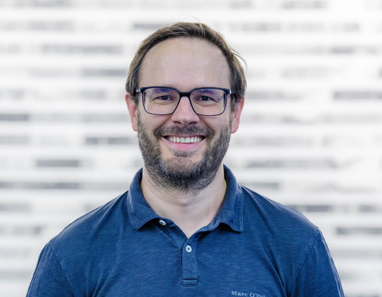 Das Portraitfoto zeigt Prof. Jens Gerken im blauen Hemd vor einem verschwommenen weißen Hintergrund mit blassen, bunten Querstreifen.