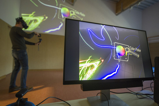 Ein Bildschirm mit farbigen Linien im Vordergrund, dahinter ein Mann mit VR-Brille und Elementen in den Händen.
