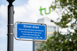 Ein blaues Straßenschild mit dem Text: "EUnet-Allee: EUnet-Projekt (European Unix NetworK) der Informatik der TU Dortmund - Dortmunder Keimzelle des deutschen Internets von 1983 -1992". Im Hintergrund verschwommen das TU-Logo auf dem Mathetower.