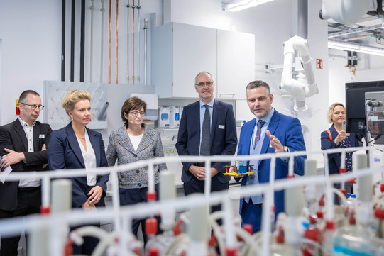 Sechs Personen stehen in einem Labor vor einem Gerät, das im unscharf im Vordergrund zu erkennen ist. Ein Mann erklärt den Anwesenden das Gerät. Unter den Personen ist Ina Brandes, Ministerin für Kultur und Wissenschaft in NRW.