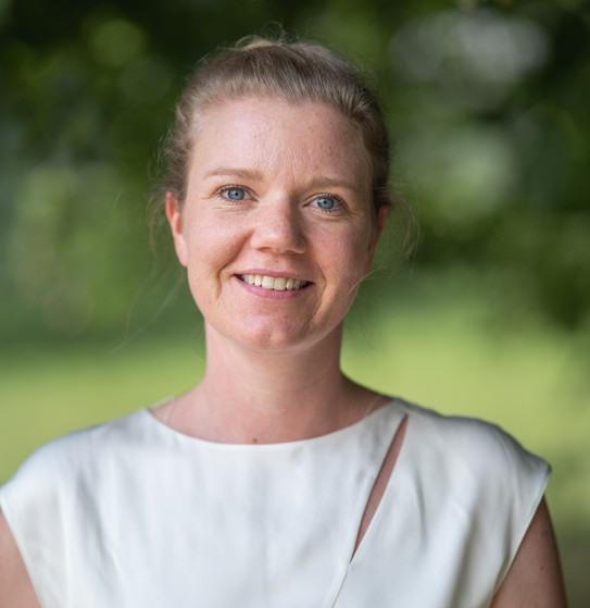 Porträtfoto von Prof. Kirsten Schorning im hochformat: Prof. Schorning trägt ein weißes Oberteil und lächelt in die Kamera. Sie steht vor einem verschwommenen grünen Hintergrund.