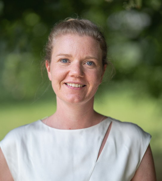 Portrait photo of Prof. Kirsten Schorning in portrait format: Prof. Schorning is wearing a white top and smiling at the camera. She is standing in front of a blurred green background.