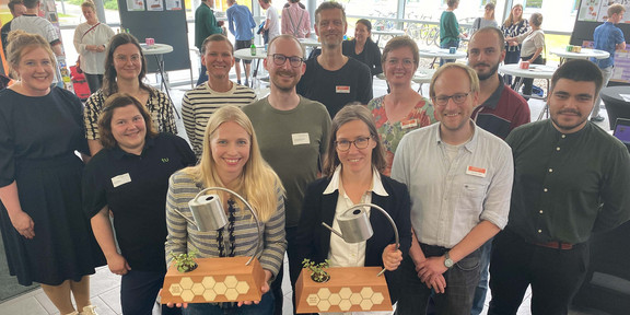 Zu sehen ist eine Gruppe von Mitarbeitenden der TU Dortmund und der FH Dortmund. Im Vordergrund stehen die Preisträgerinnen des "Impact Cups", ein Nachhaltigkeitspreis. Die Preisträgerinnen halten ihren "Pokal" in den Händen. 