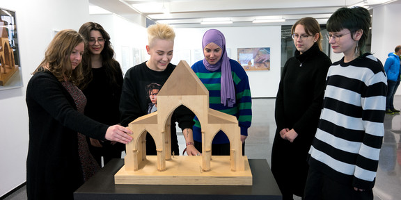 A group of young people stand around a wooden model of a building on a table.