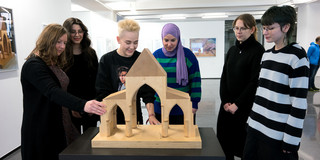 Eine Gruppe junger Menschen steht um das Holzmodell eines Gebäudes auf einem Tisch herum.