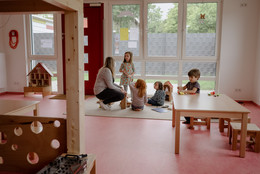 August Eine Gruppe von Kindern spielt mit einer Erzieherin auf dem Boden.