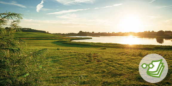 Foto: Eine grüne Landschaft mit Feldern, einem See und der untergehenden Sonne im Hintergrund.