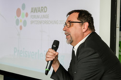 TU Sieger Junge Spitzenforschung - Laudatio Auf dem Bild ist rechts im Vordergrund ein Mann mit Mikrofon im schwarzen Anzug zu sehen mit dem Logo des Awards für Junge Spitzenforschung auf einer Leinwand im Hintergrund.