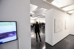 Das Bild zeigt eine durch einen Durchgang zweier Trennwände zu sehende die Auftaktveranstaltung der Ausstellung mit einer Referentin und stehendem Publikum.