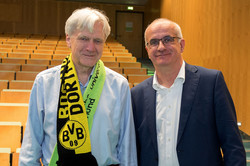 Portrait ab Taillenhöhe von Andreas von Bechholsheim (links), ein Mann mit grauen Haaren und hellblauem Hemd mit einem TU-BVB-Schal um den Hals, und Rektor Manfred Bayer (rechts), ein Mann mit dunkelblauem Jacket und Brille. Beide lächeln in die Kamera. Im Hintergrund ein leerer Hörsaal.
