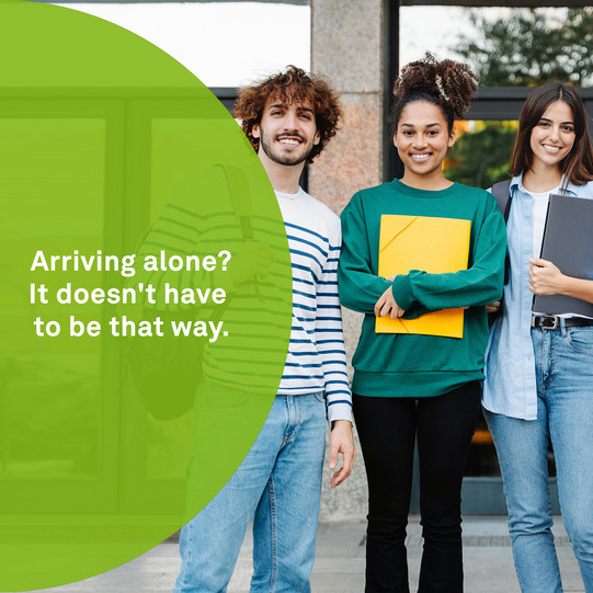 Drei junge Menschen stehen vor einem Gebäude und halten Unterlagen in den Händen. Daneben steht der Text: „Arriving alone? It doesn't have to be that way.“ – eine Botschaft, die Gemeinschaft und Unterstützung beim Ankommen vermittelt.
