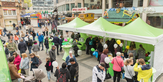 Menschen besuchen Informationsstände unter grünen Pavillons der TU in der Dortmunder Innenstadt. Die Besucher unterhalten sich, halten Luftballons in den Händen und informieren sich über verschiedene Angebote der TU Dortmund. Im Hintergrund sind weitere Marktstände und Gebäude zu sehen.