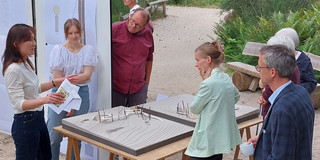 6 Personen stehen in einer Parkanlage auf einem sandigen Boden um einen Tisch mit zwei Modellen eines Steges. Hinter dem Tisch stehen zwei Pinnwände an denen verschiede Zeichnungen angepinnt sind.