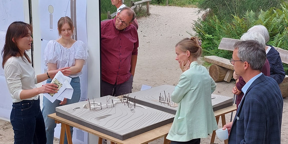 Präsentation des Steges für den Rombergpark 6 Personen stehen in einer Parkanlage auf einem sandigen Boden um einen Tisch mit zwei Modellen eines Steges. Hinter dem Tisch stehen zwei Pinnwände an denen verschiede Zeichnungen angepinnt sind.