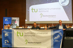 Drei Männer halten einen Banner der TU Dortmund in den Händen. Der Kanzler hält außerdem einen Preis in der Hand.
