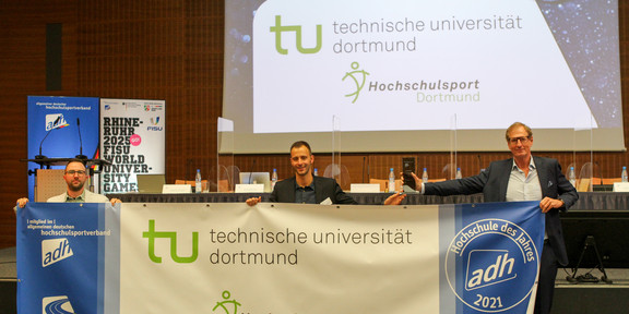 Drei Männer halten einen Banner der TU Dortmund in den Händen. Der Kanzler hält außerdem einen Preis in der Hand.