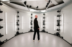 Das Bild zeigt eine schwarz gekleidete, männliche Person in der Mitte eines weißen, ründlichen Raumes und dreht seinen Oberkörper nach Hinten, um in die Kamera zu lächeln. An der Wand und Decke hängen Kameras in gleichmäßigen Abständen übereinander.