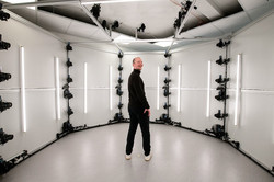 Das Bild zeigt eine schwarz gekleidete, männliche Person in der Mitte eines weißen, ründlichen Raumes und dreht seinen Oberkörper nach Hinten, um in die Kamera zu lächeln. An der Wand und Decke hängen Kameras in gleichmäßigen Abständen übereinander.