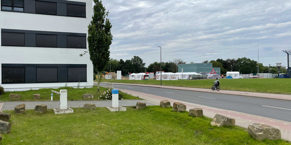 Auf der gegenüberliegenden Seite einer Straße sieht man rechts auf einer Wiese mehrere Zelte. Links auf der dem Betrachter näheren Straßenseite ein weißes Bürogebäude mit der orangenen Aufschrift „FH Dortmund“. Davor Parkplätze und eine Wiese.