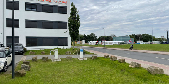 On the opposite side of a road, you can see several tents on a meadow on the right. To the left, on the side of the road closer to the viewer, is a white office building with the orange inscription "FH Dortmund". In front of it are parking lots and a meadow.