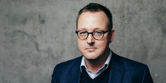 Portrait Philipp Grollmann Portrait eines Mannes mit Anzug und Brille vor einer grauen Wand