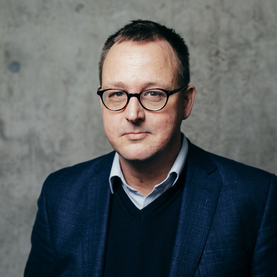 Portrait eines Mannes mit Anzug und Brille vor einer grauen Wand