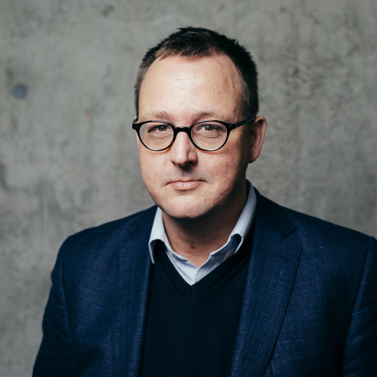 Portrait Philipp Grollmann Portrait of a man wearing glasses and a suit in front of a gray wall