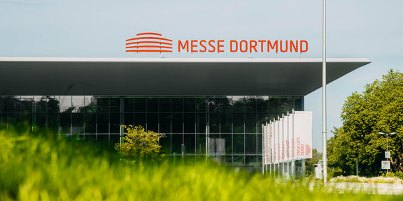 Aufnahme eines großen Gebäudes, auf dem Dach die Aufschrift "Messe Dortmund". Im Vordergrund ist verschwommen eine Wiese zu sehen.