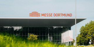 Messe Dortmund Aufnahme eines großen Gebäudes, auf dem Dach die Aufschrift "Messe Dortmund". Im Vordergrund ist verschwommen eine Wiese zu sehen.