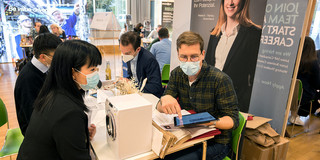 Miele information stand at the International Career Fair Two men are sitting at a table in front of a Miele display. One of the men is showing something on his tablet to a woman sitting opposite him. Between them is a miniature washing machine. Other information stands can be seen in the background.