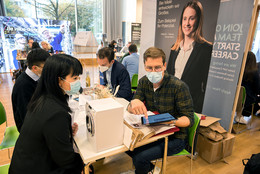 Zwei Männer sitzen vor einem Aufsteller von der Firma Miele an einem Tisch. Einer der Männer zeigt einer ihm gegenüber sitzenden Frau etwas auf seinem Tablet. Zwischen den beiden steht eine Miniaturwaschmaschine. Im Hintergrund sind weitere Infostände zu sehen. 