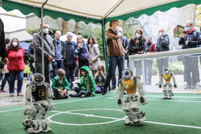  Mehrere Personen stehen um ein kleines grünes Fußballfeld herum, auf dem drei kleine weiße Roboter spielen.