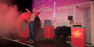 Marcus Weber Ein Mann steht jubelnd auf einer Bühne, aus einem Apparat kommt Rauch