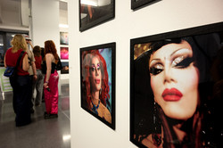 Nahaufnahme von zwei Kunstwerken in einer Galerie. Die Kunstwerke zeigen Drag Queens. Im Hintergrund stehen Gäste der Galerie.