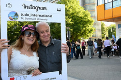 Eine Frau und ein Mann halten gemeinsam einen Fotorahmen, der wie ein Instagram-Post gestaltet ist, und lächeln. Im Hintergrund sind Campusbesucher*innen zu sehen.
