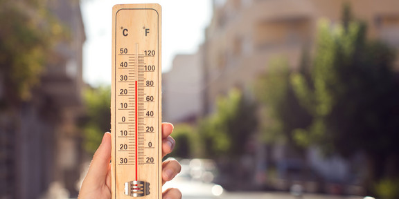 Hitze in der Stadt Eine Hand hält ein Thermometer, das etwa 30 Grad Celsius anzeigt. Im Hintergrund verschwommen eine Stadt im Sommer.