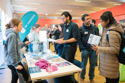 An einem Karrieremessestand stehen rechts zwei internationale Studenten und eine internationale Studentin, die sich Flyer ansehen. Auf dem Tisch liegen verschiedene Materialien der Firma. Links hinter dem Tisch steht eine Firmenvertreterin.