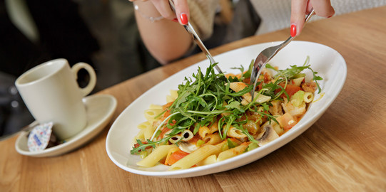 food A plate with pasta and a cup are standing on a table. Hands are cutting the food.
