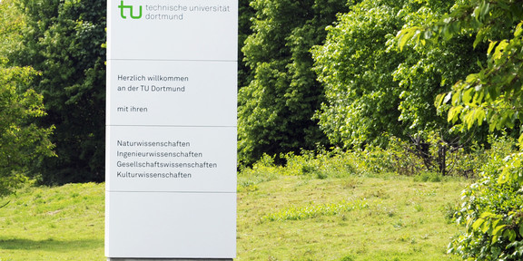 TU Dortmund Ein großes weißes Schild mit dem TU-Logo und dem Schriftzug "Herzlich Willkommen an der TU Dortmund" steht auf einer Wiese mit Bäumen im Hintergrund.