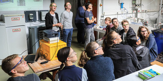 SchnupperUni Eine Gruppe Schüler*innen sitzt in einem Halbkreis vor drei präsentierenden Personen und einem Beamer. Alle Personen im Bild betrachten etwas außerhalb des rechten Bildrandes.